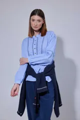 Camisa blanca de algodón con pechera decorada con alforzas y puntillas. Presenta cierre frontal con botones, mangas tres cuartos con frunces y detalles de puntilla, y un diseño de corte holgado con ruedo redondeado.