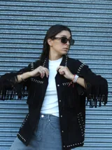 Chaqueta de gamuza negra con flecos en el pecho y mangas, decorada con tachas metálicas en el canesú, cuello y ruedo. Posee cierre frontal y forro interior.