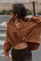 Camisa de lino color beige, con cuello solapa, escote en V y mangas largas. Presenta un diseño de bajo asimétrico con pliegues delanteros que generan un efecto abullonado.