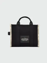 Bolso tipo tote de lona color negro con letras blancas bordadas en el frente que dicen 'THE TOTE BAG MARC JACOBS'. Cuenta con asas superiores cortas y una correa de hombro ajustable y extraíble.
