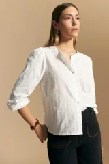 Camisa de algodón blanca con delicadas líneas bordadas verticales y textura calada. Presenta escote redondo con cierre frontal de botones, mangas largas con leve volumen en los hombros y puños elásticos.