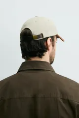 Gorra de seis paneles de algodón en color beige con visera marrón contrastante. Cuenta con un diseño bordado en marrón en el frente con texto alusivo.