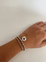 Set de dos pulseras plateadas elásticas con cuentas redondas y dije circular con una cruz calada.