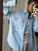 Camisa oversize de corte recto, confeccionada en tela de algodón a rayas verticales celestes y blancas. Presenta cuello clásico, cierre frontal con botones y mangas largas. El diseño destaca por una estampa frontal con palabras relacionadas al amor en diversos idiomas en color bordó.