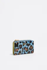 Billetera mediana con exterior rígido y textura brillante, con estampado de leopardo en tonos azul celeste, negro y naranja. Presenta cierre de cremallera y logo metálico de Bimba y Lola en el frente.