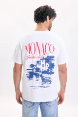 Remera blanca de manga corta con gran estampado en la espalda que incluye la inscripción 'Monaco Yacht club' en rojo y una ilustración azul de un paisaje costero.