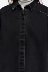 Camisa de corte oversize confeccionada en mezcla de algodón, con cuello camisero, cierre frontal de botones y manga larga. Presenta detalles de remaches metálicos a lo largo de las costuras de las mangas y el cuello.