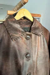Tapado de cuero marrón con diseño artesanal, cuello camisero con detalles de pasamanería en gamuza, cierre frontal con botones de guampa y bolsillos delanteros con bordes decorativos. Presenta un corte cómodo y versátil.