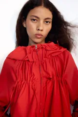Chaqueta corta de nylon color beige, con cuello subido, capucha y cierre frontal con cremallera. Presenta mangas largas con puños elásticos, bolsillos laterales y detalles de cordones ajustables con lazadas en el pecho y el bajo.