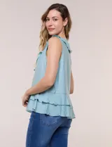 Blusa blanca sin mangas confeccionada en tejido de satén con suave textura arrugada y caída fluida. Cuenta con escote en V con volante en el cuello y ruedo estilo peplum con doble capa de volados superpuestos.