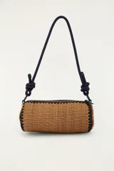 Bolso tipo bandolera de formato cilíndrico, confeccionado con material trenzado en color tostado, con detalle de bordado frontal que dice 'Rio' acompañado de una palmera y un tucán. Cuenta con una correa larga de cuerda oscura con nudos ajustables y cierre superior.