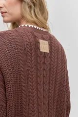 Cardigan de punto con diseño de trenzas, color marrón, con cierre frontal de botones y detalle de borde en color blanco. Presenta bolsillos frontales y mangas largas.