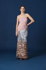 Vestido largo de corte ajustado con breteles finos y escote recto. Presenta un diseño estampado que combina un fondo rosado en la parte superior con una ilustración paisajística de montañas, bosques y ciervos en tonos grises y marrones en la parte inferior.