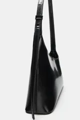 Bolso de hombro de diseño asimétrico en color negro, con acabado brillante de charol, asa ajustable y cierre de cremallera.