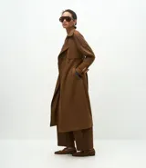 Trench coat largo de gamuza sintética con cuello peaked, cierre frontal cruzado con botones y cinturón ajustable en la cintura. Presenta mangas largas con reguladores en los puños y forro interior.