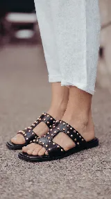 Sandalias planas de cuero negro, con diseño de tiras anchas interconectadas y punta cuadrada, decoradas con tachas metálicas doradas.
