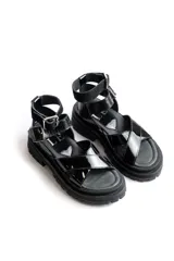 Sandalias negras de estilo chunky con tiras cruzadas en charol y base de goma dentada. Cuentan con una tira superior desmontable y cierre con hebilla metálica en el tobillo.