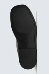 Bota corta de piel negra, estilo motociclista, con caña media y punta cuadrada. Presenta una correa ajustable con hebilla metálica en el tobillo y otra en la parte superior de la caña.