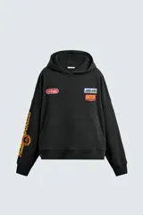 Sudadera tipo hoodie de corte relajado, color negro, con capucha y bolsillo frontal tipo canguro. Presenta parches decorativos en el pecho y estampados en color amarillo en la manga derecha. Acabados en rib en puños y ruedo.