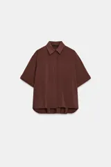 Camisa de cuello solapa y manga corta con dobladillo. Presenta un diseño de corte holgado con bajo asimétrico y aberturas laterales. Cierre frontal mediante botones ocultos bajo una tapeta.