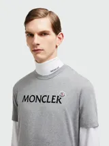 Remera de algodón gris con cuello redondo y logo de la marca estampado en el pecho junto con un pequeño parche circular.