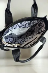 Cartera de cuero modelo Tali, de diseño estructurado que combina los colores blanco, negro y dorado, con detalles de arte pintado a mano en el frente. Cuenta con doble asa de mano, correa larga desmontable y herrajes metálicos.