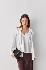 Camisa blanca de manga larga con textura suave y caída liviana, cuello camisero, puños anchos y fruncidos sutiles en los hombros.