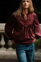 Blusa de satén color bordó con cuello redondo, cierre frontal con cordones y detalle de canesú con bordado calado y volados. Presenta mangas largas abullonadas con puños elásticos.