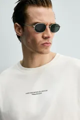 Camiseta de corte relajado, color crudo, con cuello redondo y manga corta. Presenta un diseño con texto estampado y bordados a contraste en la parte frontal.