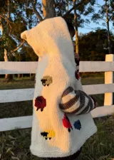 Poncho tejido de lana color crema con capucha y mangas a rayas horizontales en tonos grises. Presenta bordados de ovejas de colores en el frente.