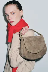 Bolso bandolera de piel gamuzada color gris topo, con solapa curva, asa de mano corta y herrajes metálicos dorados.
