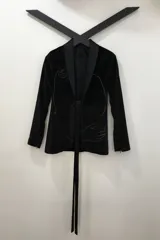 Blazer tipo smoking de terciopelo negro con acabado brillante y detalles estampados en dorado. Presenta solapas clásicas, cierre frontal cruzado mediante un lazo y mangas largas con cierres incorporados. El diseño es entallado y sofisticado.