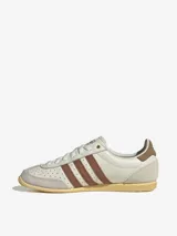 Championes Adidas modelo Japan, con diseño retro de perfil bajo. Presentan un cuerpo de cuero color crema con perforaciones en la puntera, icónicas tres tiras laterales en color marrón y refuerzos de gamuza gris en la puntera y el talón. Incluye suela de goma color amarillento y detalles dorados en el lateral y la lengüeta.