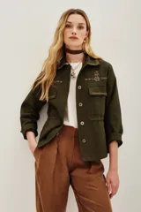 Campera estilo militar o safari color verde oliva, confeccionada en algodón. Presenta cuello solapa y bolsillos frontales con tapa. Destaca por sus apliques de pedrería o bordados en tonos marrones y dorados en el cuello y sobre los bolsillos.