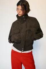 Cazadora tipo bomber de tejido efecto ante color marrón oscuro, con cuello subido, manga larga y cierre frontal con cremallera y botones ocultos. Presenta bolsillos frontales con solapa y botones metálicos.