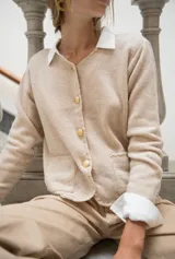 Cardigan de punto color beige con cuello redondo, cierre frontal con botones dorados, bolsillos frontales y terminaciones en crochet. Presenta hombreras internas y diseño de corte recto.