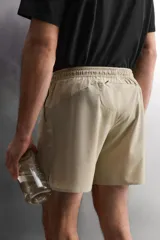 Pantalón corto deportivo color marrón oliva, confeccionado en tejido técnico ligero y elástico, con forro interior. Presenta cintura elástica ajustable con cordón, bolsillos frontales, bolsillo de seguridad trasero con cierre y aberturas laterales en el bajo.