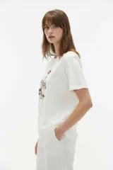 Remera de manga corta color blanco, de corte holgado con cuello redondo a la base y estampado frontal en tono bordó de un gato y un ovni en un paisaje desértico.
