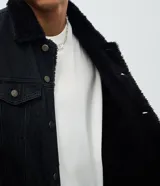 Chaqueta de jean color negro con cuello de sherpa, cierre frontal con botones metálicos, bolsillos con solapa en el pecho y bolsillos laterales. Diseño de corte regular y manga larga.
