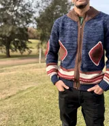 Campera de punto con cierre frontal, cuello alto y diseño artesanal. Presenta un cuerpo en tono azul jaspeado con detalles en marrón, blanco y rojo, incluyendo parches en forma de rombo en el pecho y los codos.