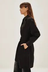 Trench negro de corte midi con cuello camisero, cierre frontal y cinturón ajustable a la cintura. Presenta mangas largas con puños rectos y un diseño minimalista y versátil.