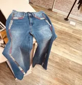 Jean de corte recto y tiro medio, confeccionado en denim azul con lavado desgastado. Presenta detalles de bordado artesanal en hilo de colores a lo largo de las costuras laterales y en los bolsillos delanteros.