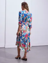 Vestido midi con estampado floral en tonos azules, rojos y blancos. Tiene escote en V, mangas 3/4 con puños fruncidos, cintura elastizada con lazo de ajuste y falda con volantes.