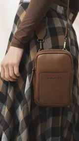 Bandolera pequeña de diseño compacto en color mostaza, confeccionada en material sintético con textura granulada. Presenta un bolsillo frontal con el logo de la marca impreso, cierre superior y correa ancha ajustable y desmontable.