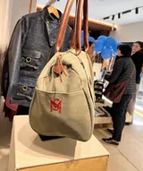 Bolso amplio de gamuza azul con manijas largas de cuero marrón. Cuenta con bolsillos laterales, uno con clip, cierre posterior y un bolsillo interno amplio. Presenta detalle de bandera y logo bordado en los laterales.