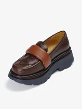 Mocasín de plataforma con capellada de cuero marrón oscuro trenzado y banda superior acolchada de cuero color caramelo. Posee una suela robusta de goma con capas en tonos marrón y azul marino.