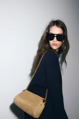 Bolso de hombro confeccionado en piel de gamuza color marrón, con diseño estructurado y pespuntes decorativos en la solapa. Cuenta con una correa de cadena metálica dorada con refuerzo de piel y cierre imantado.