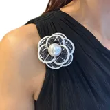 Broche en forma de flor tipo camelia, confeccionado en tela de tweed gris con bordes en cordón blanco y una perla central. Cuenta con un gancho versátil para sujetar en prendas o en el cabello.