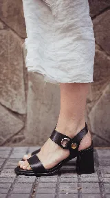Sandalias de cuero color negro con taco cuadrado, tira ancha en el empeine y tira al tobillo con detalle de argolla y botón metálico dorado.