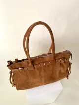 Bolso grande de cuero marrón con textura tipo cocodrilo, estructura rígida y asas para llevar al hombro. Tiene una tira con tachas y hebillas en cada extremo, también lleva lazos en los costados.